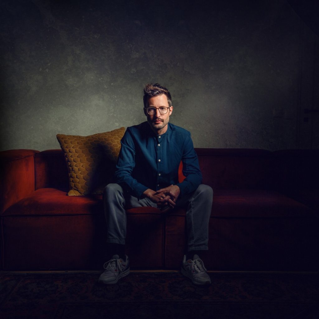 Editorial photo of composer Roy Bemelmans sitting on a couch. Deep colors.